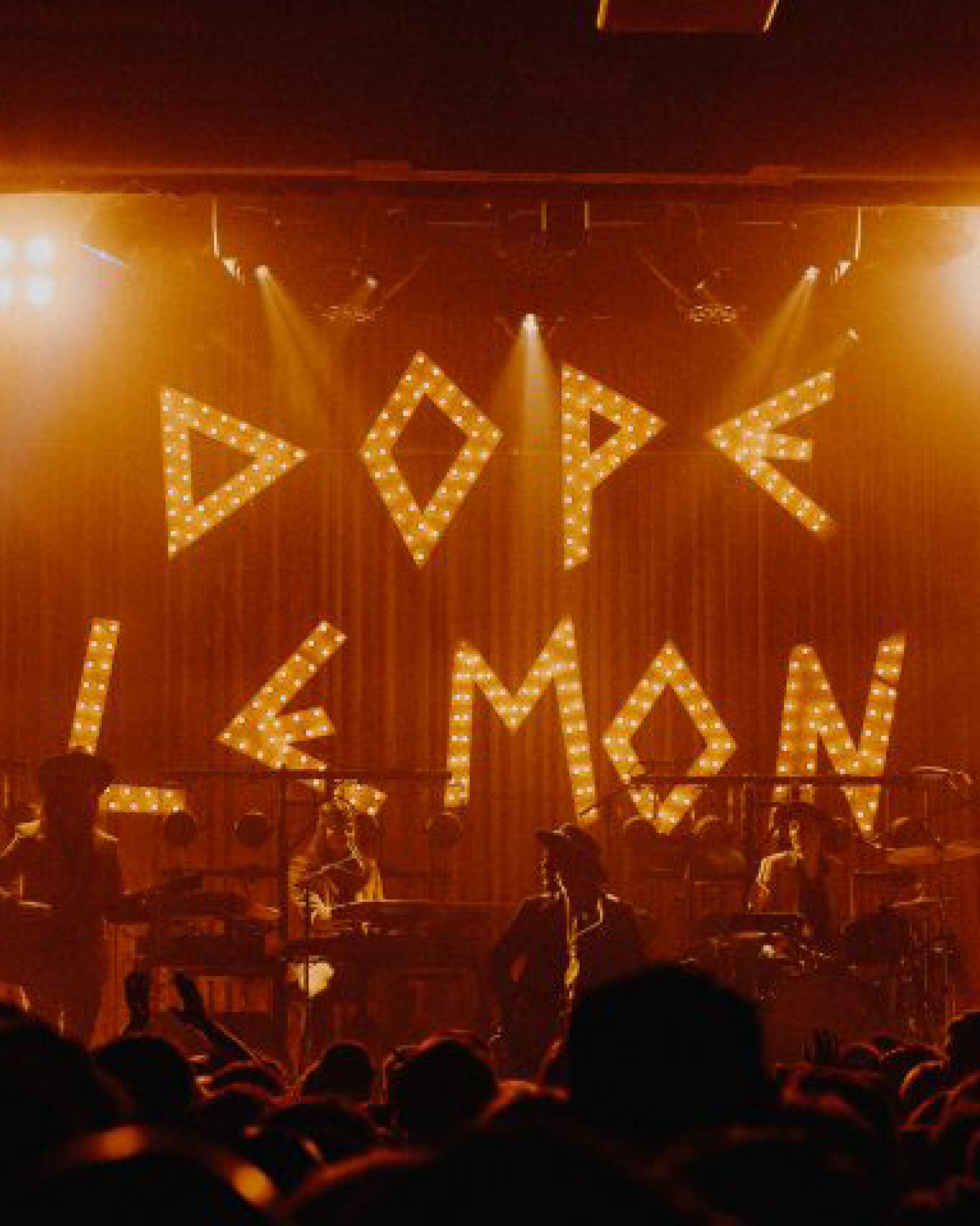 Live Dope Lemon concert scene with illuminated marquee letters spelling "DOPE LEMON" in bold lights on stage. Silhouettes of the band and crowd create a golden, dreamlike atmosphere—capturing the spirit of the FBS x Dope Lemon collaboration.