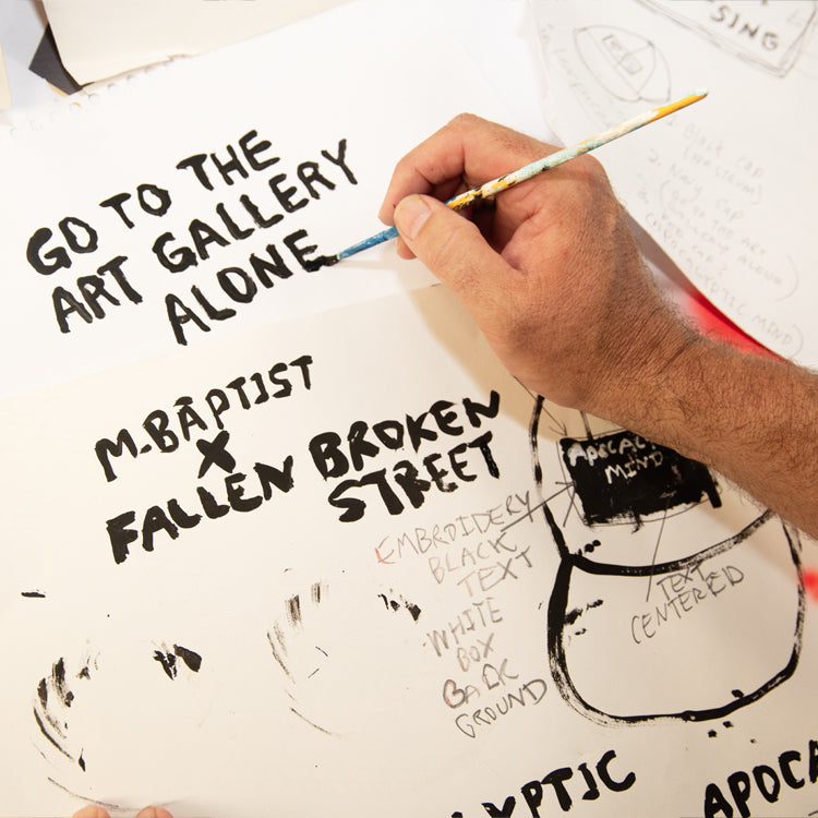 Artist Marty Baptist paints “GO TO THE ART GALLERY ALONE” alongside “M. BAPTIST x FALLEN BROKEN STREET” in black text. A behind-the-scenes look at the creative process behind the Fallenbrokenstreet x Marty Baptist collaboration.