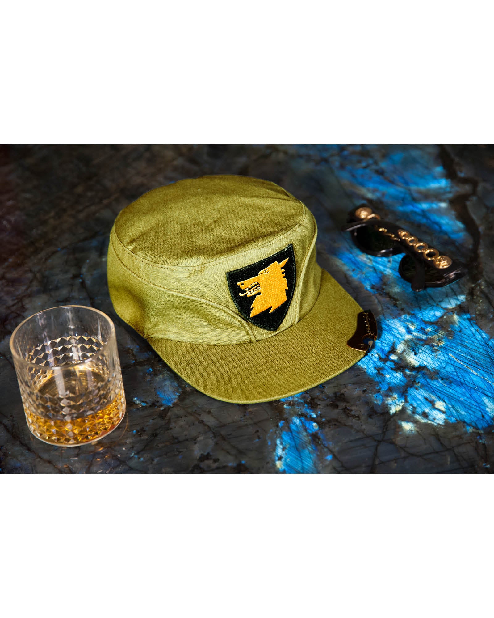 A canvas cap in green color with a brass bottle opener on the brim, accompanied by a glass of whiskey and a bottle, against a backdrop with a blue and black pattern.