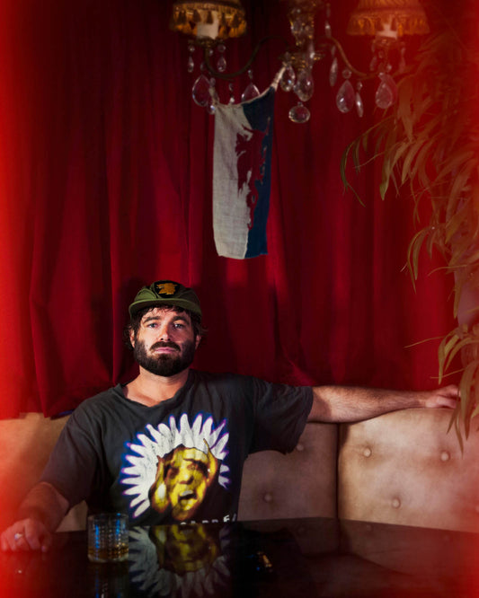 Australian artist Angus Stone wearing the Wolf Pack Cap by FallenBROKENstreet, photographed against a deep red curtain backdrop. Canvas cap in olive with wolf patch, featuring patent brass bottle opener brim. Styled with a casual t-shirt, showcasing the collaboration’s rugged yet artistic edge.