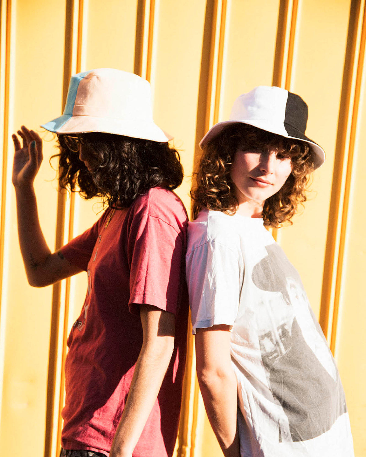 Two women wear fallen broken street bucket hats, the hats are two-toned in colour. They are standing back to back with a relaxed street style