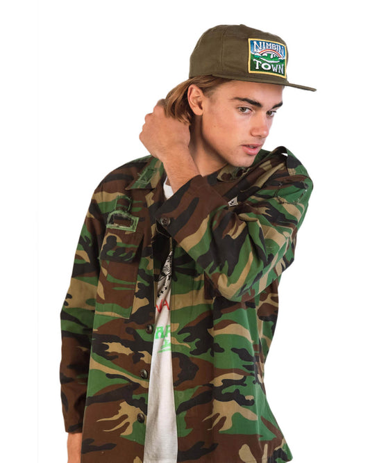 Model styles The Stash Cap – Canvas Nimbin with a camo shirt and effortless attitude. Features embroidered patch, structured fit, and secret stash pocket for hidden essentials.