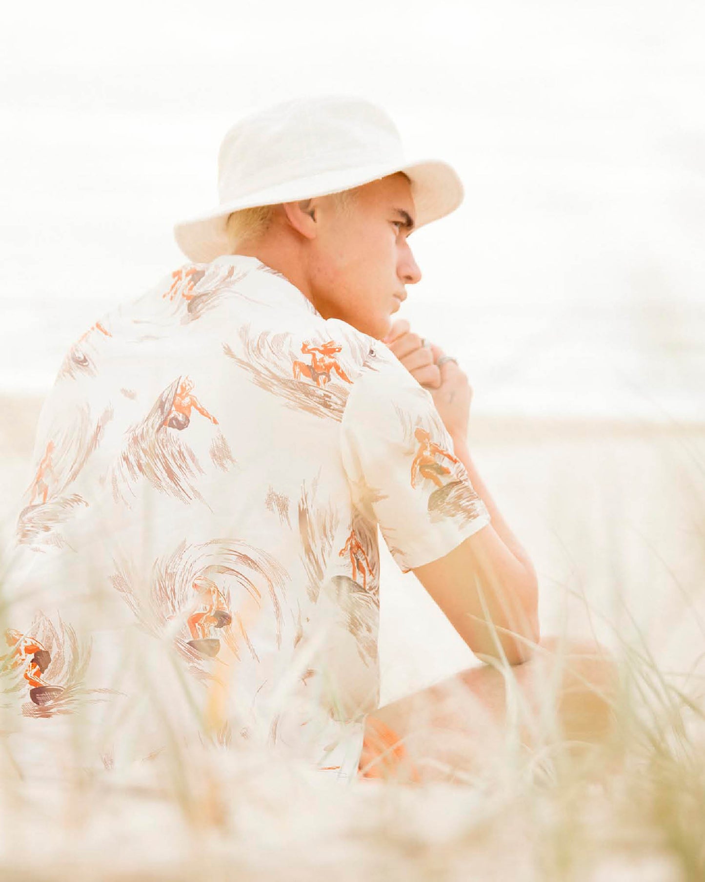 Model wearing the Saturday Bucket Hat – White by FallenBROKENstreet. Cotton chenille wide brim hat with UPF 50+ sun protection, styled with a floral shirt and bright shorts. Photographed outdoors at the beach, highlighting casual unisex summer styling.