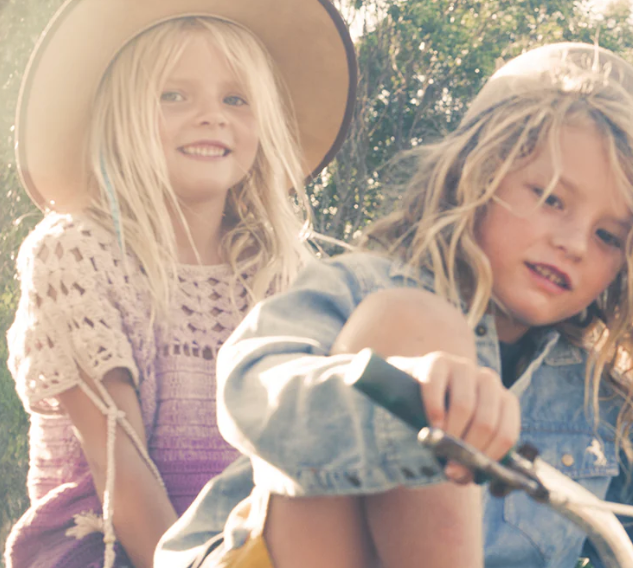 two kids ride a bike in the summer wearing fallen broken street kids hats.