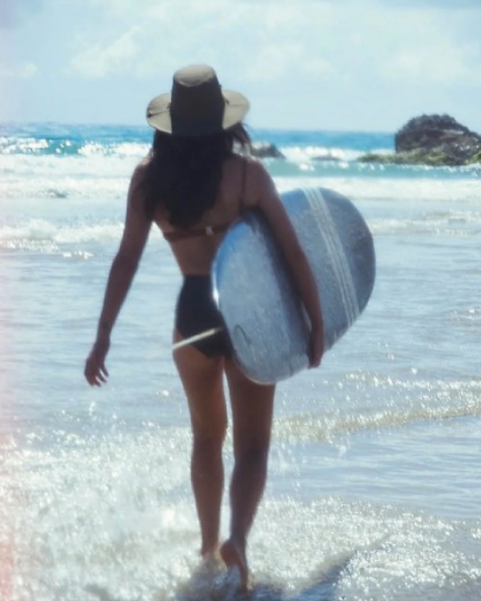 A women walks up the beach in Byron Bay with a long board, she is wearing a fallen broken street surf hat.