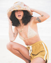 Model wearing the Sunday Bucket Hat – Paisley by FallenBROKENstreet. Lightweight cotton canvas bucket hat with wide brim and all-over paisley print. Styled with a crochet bikini top and shorts at the beach, highlighting its unisex, travel-ready design made for sun protection and coastal adventures.