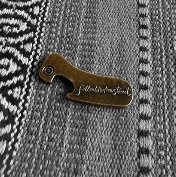 Fallenbrokenstreet brass bottle opener photographed on textured fabric. Compact 5cm x 2cm design with embossed Fallenbrokenstreet logo, crafted from solid brass. A timeless accessory for festivals, travel, and home.