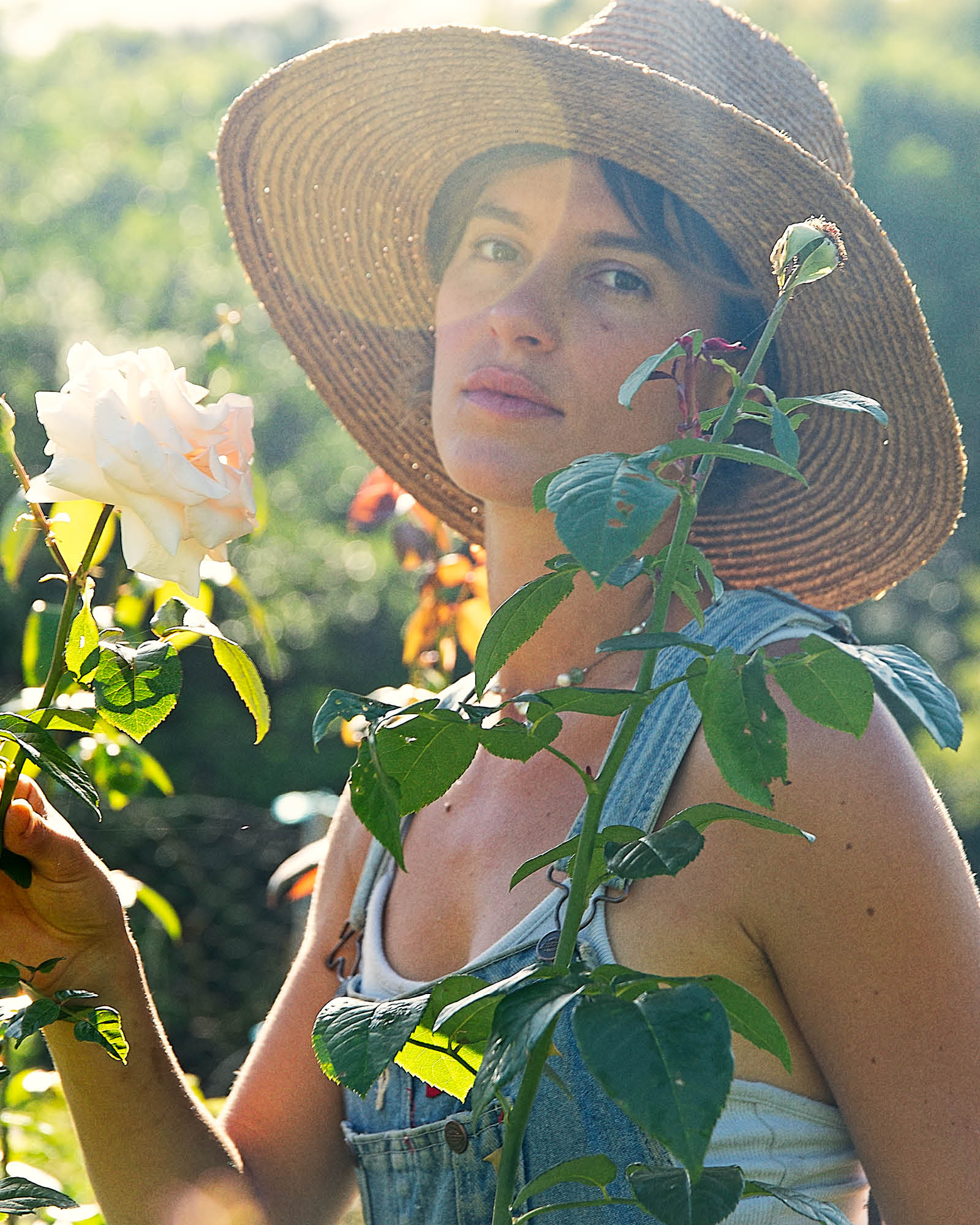 Woman wears the Fallenbrokenstreet straw hat in a lush garden, surrounded by blooming leaves and dappled light. The hat’s feather and ribbon details add vintage charm.