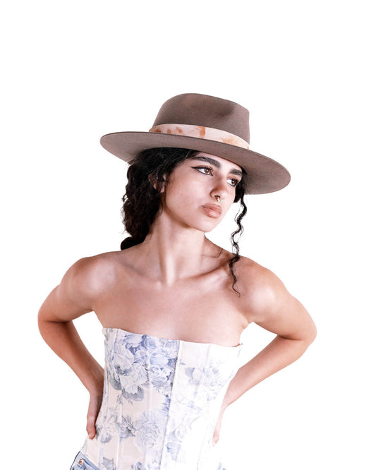 A woman models The Zephyr felt hat in vintage brown against a white backdrop. Styled with a strapless floral dress, the image highlights the hat’s bold brim, frayed cotton band, and hand-shaped crown. This wide-brimmed wool hat offers both elegance and function, making it the perfect accessory for summer adventures or road trips. FallenBROKENstreet signature style with vintage flair.