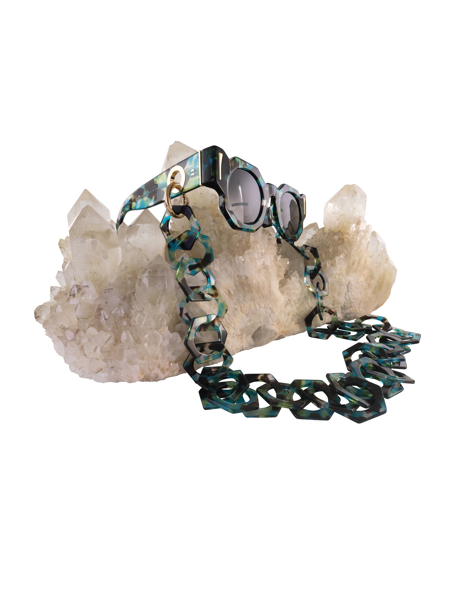 Close-up of THE COSMOS – Galaxy/Black sunglasses hanging off quartz crystal, showcasing chain compatibility, acetate pattern, and gold hardware.