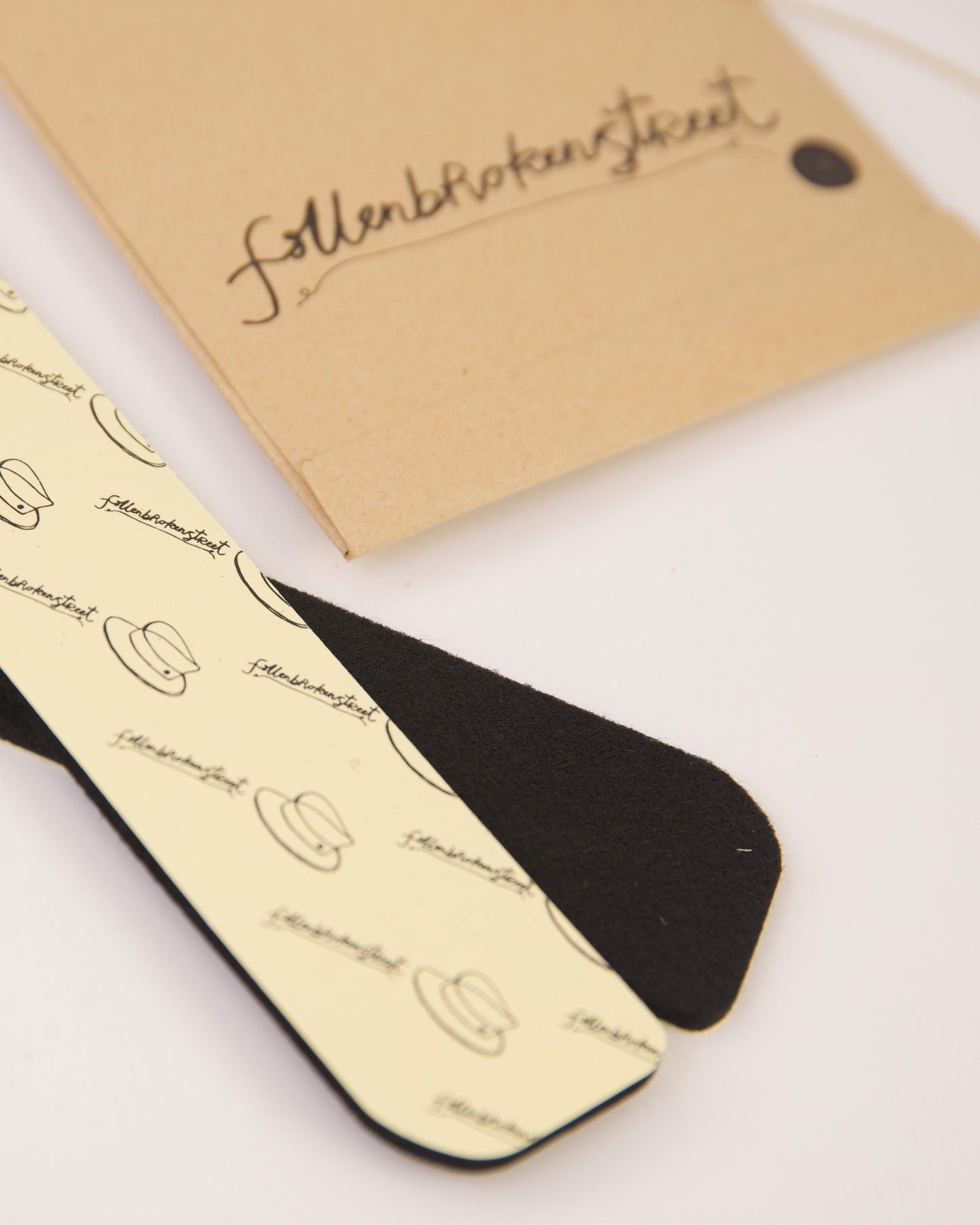 Fallenbrokenstreet hat filler strips displayed with branded kraft tag packaging. Includes one black and one printed cream felt strip with adhesive backing. Perfect for making your favourite hat fit just right, these strips reduce the inner circumference by up to 2.5cm. Designed to be used with wool felt or straw hats, the fillers are easy to apply and remove. Great for styling flexibility and hat stability. The branded packaging adds an artisanal touch, aligning with Fallenbrokenstreet's thoughtful accessor