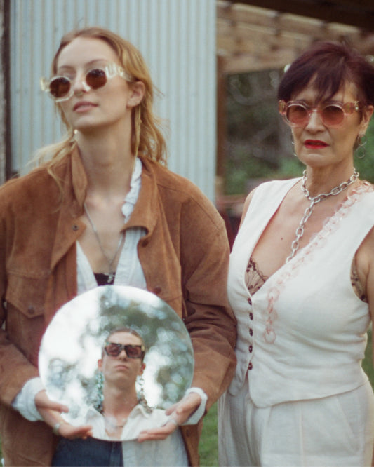 Editorial lifestyle photo of two women wearing THE COSMOS – Pink/Pink sunglasses. Retro styling, standing outdoors, one holding a mirror with a man’s reflection. Playful and cinematic vibe.
