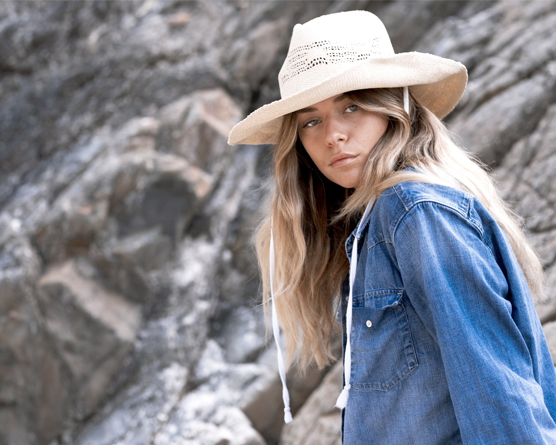Model perched on rocky terrain wearing The Lover Straw Hat – Natural. This shot highlights the hat’s flexibility and comfort in real-world adventures, from hikes to beachside wandering. The natural tones blend beautifully with the rugged backdrop.