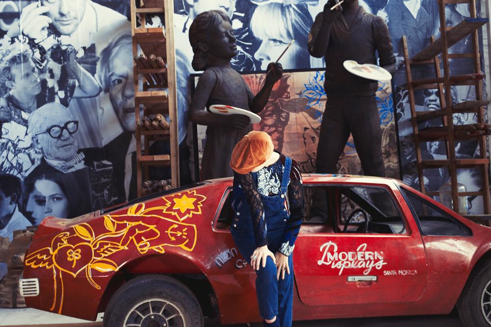The Bandit Paperboy Cap – Tan Corduroy by FallenBROKENstreet. Corduroy construction, leather strap, and metal button detail. Designed for comfort, attitude, and lasting style shown on a girl in front of a vintage car with david bromley's paintings and sculpture
