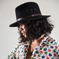 Person wearing a black hat with a brown band and a colorful floral shirt on a plain background