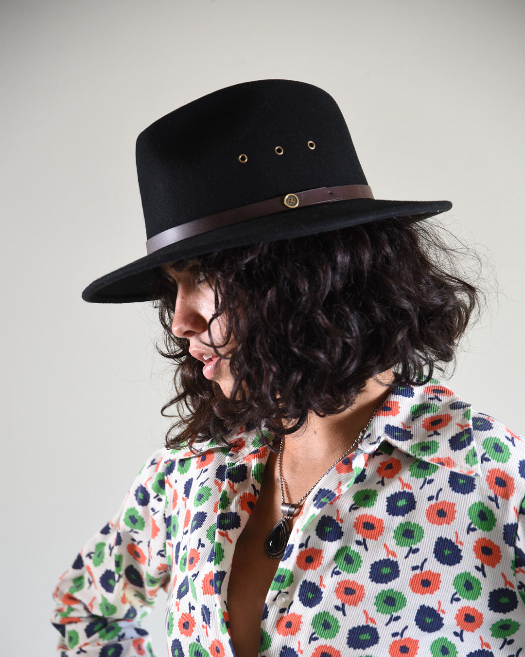 Person wearing a black hat with a brown band and a colorful floral shirt on a plain background