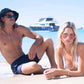 Two models sitting together in swimwear wearing Dope Lemon sunglasses with a yacht behind them.