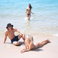 Two models sitting on the beach in sunglasses and swimwear, ocean in the background.