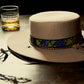 Lifestyle image of the Troubadour Felt Hat – FallenBROKENstreet x Angus Stone. Wool felt hat with custom ribbon and braided strap, placed on a wooden table with a glass of whiskey and chain. Evokes storytelling, adventure, and the spirit of the travelling musician.