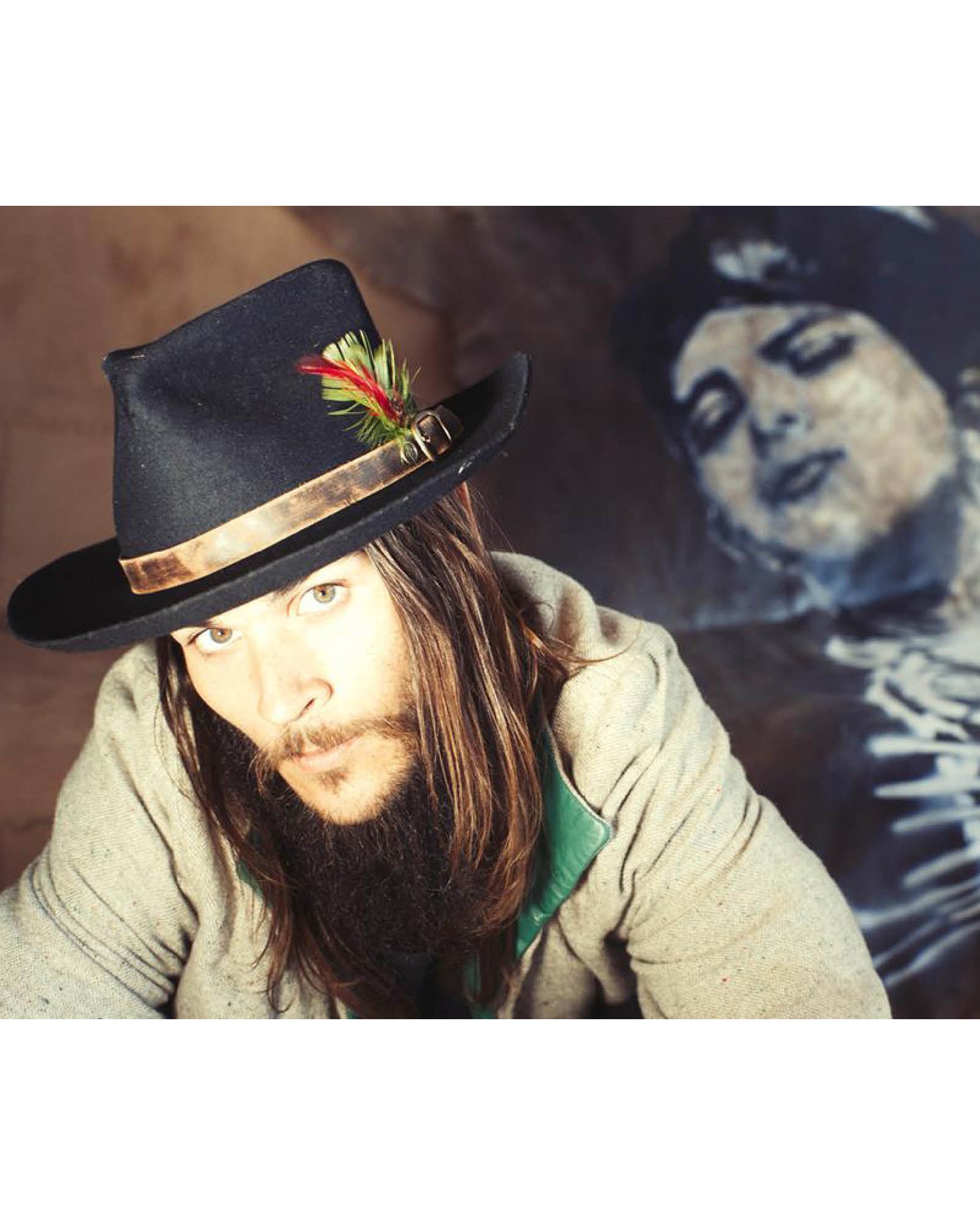 Man wearing a black felt hat with a leather band and feather detail from the FBS x David Bromley collection. A painted portrait looms in the background, blending fashion and fine art in this moody, expressive campaign shot by Fallenbrokenstreet.