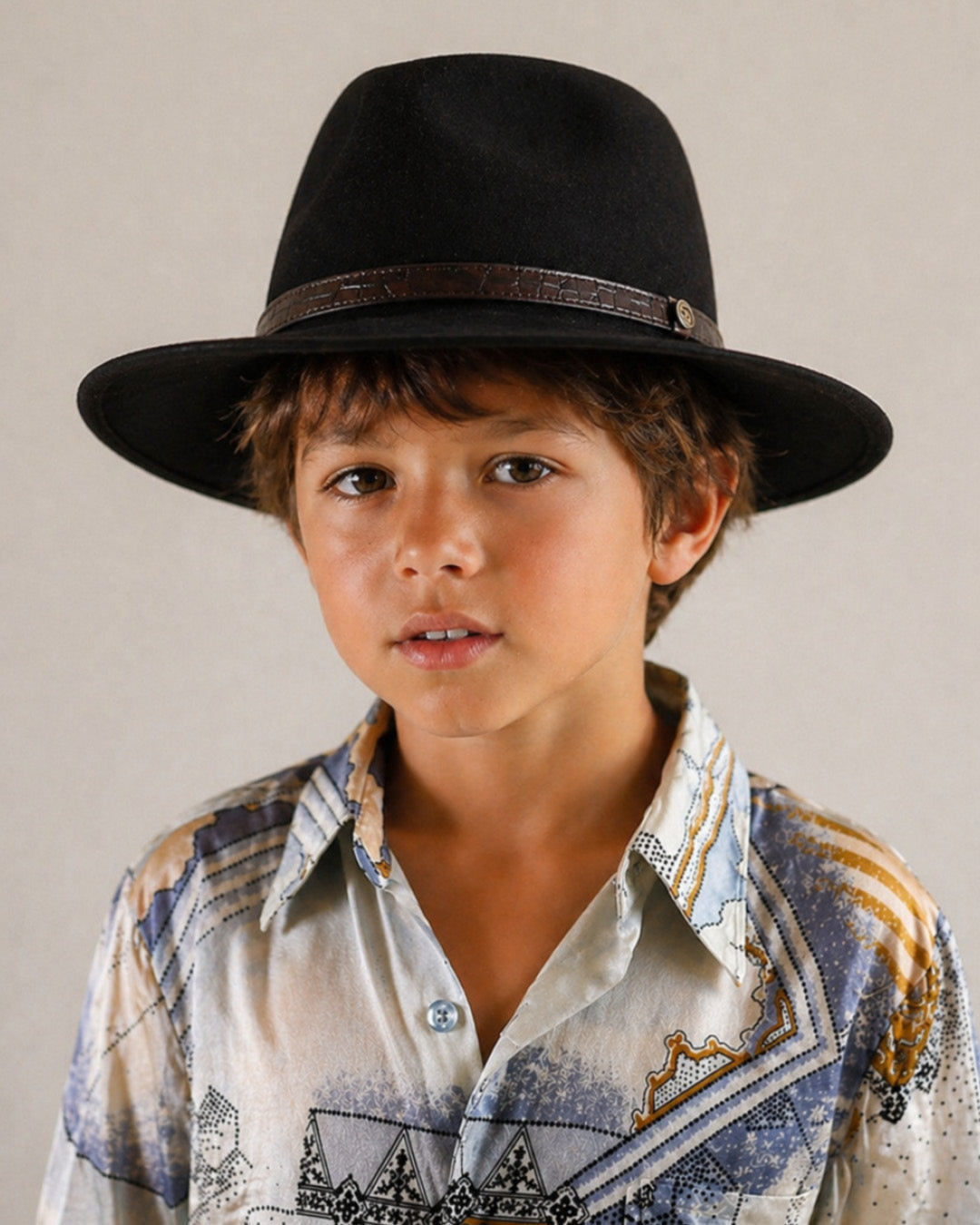 Child wearing a black fedora hat and patterned shirt against a neutral background