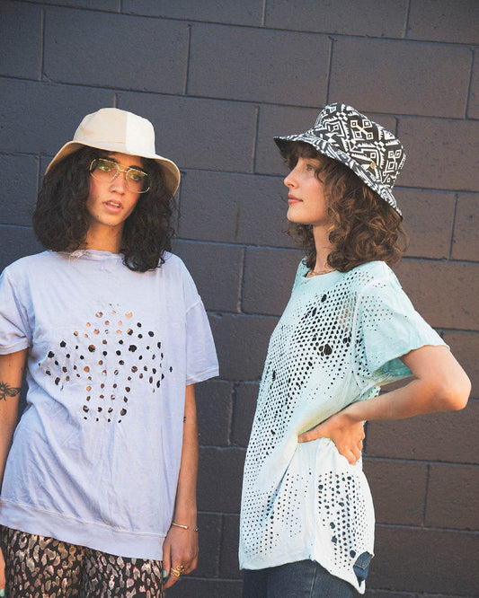 Models wearing the FlipSide Bucket Hat – Aztec Reversible by FallenBROKENstreet. One shows the bold Aztec patterned side, the other the plain reverse. Cotton canvas bucket hat with wide brim for sun protection, styled with relaxed summer clothing against a colourful wall backdrop.