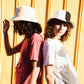 Two women wear fallen broken street bucket hats, the hats are two-toned in colour. They are standing back to back with a relaxed street style