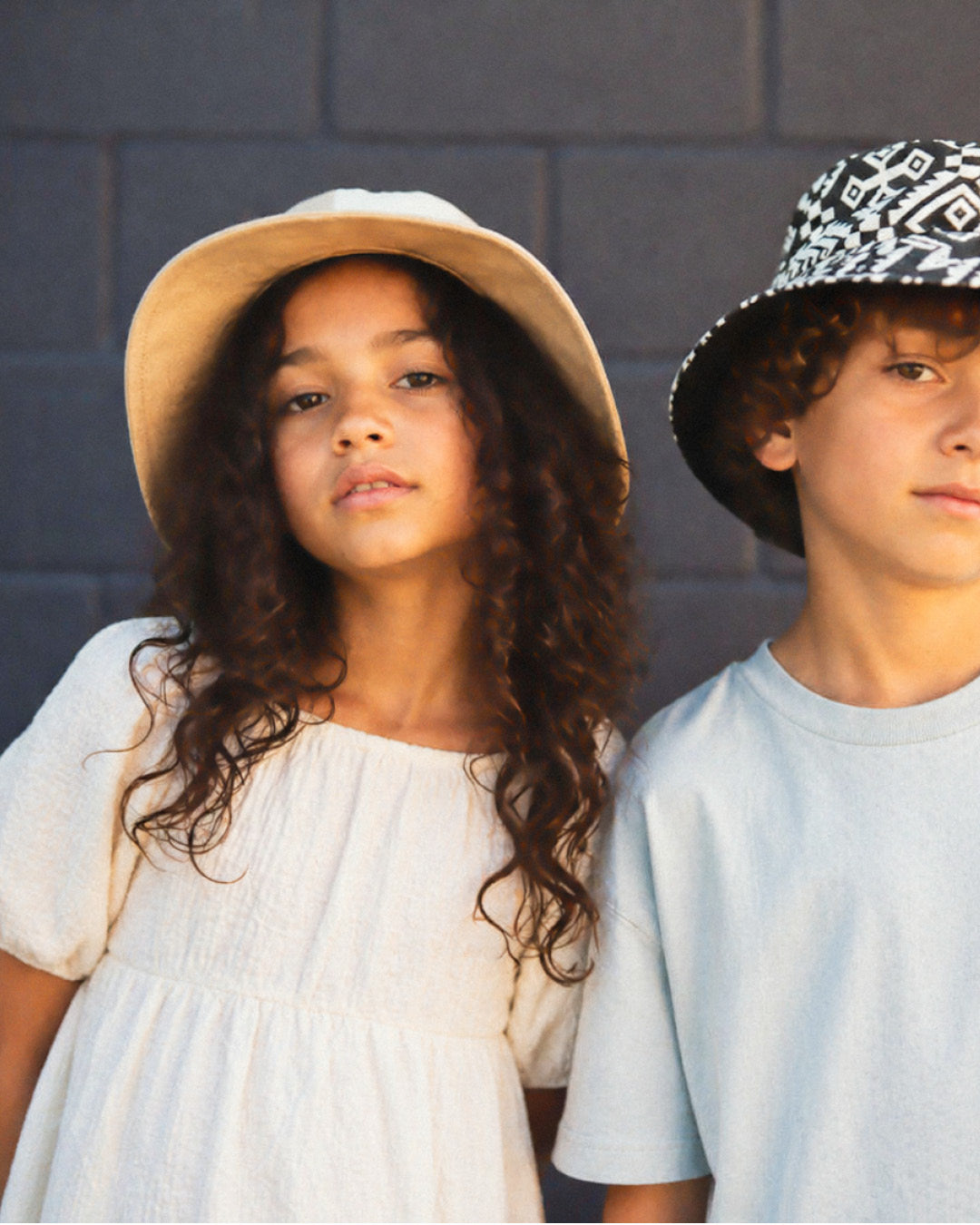 Two children wearing hats against a gray wall