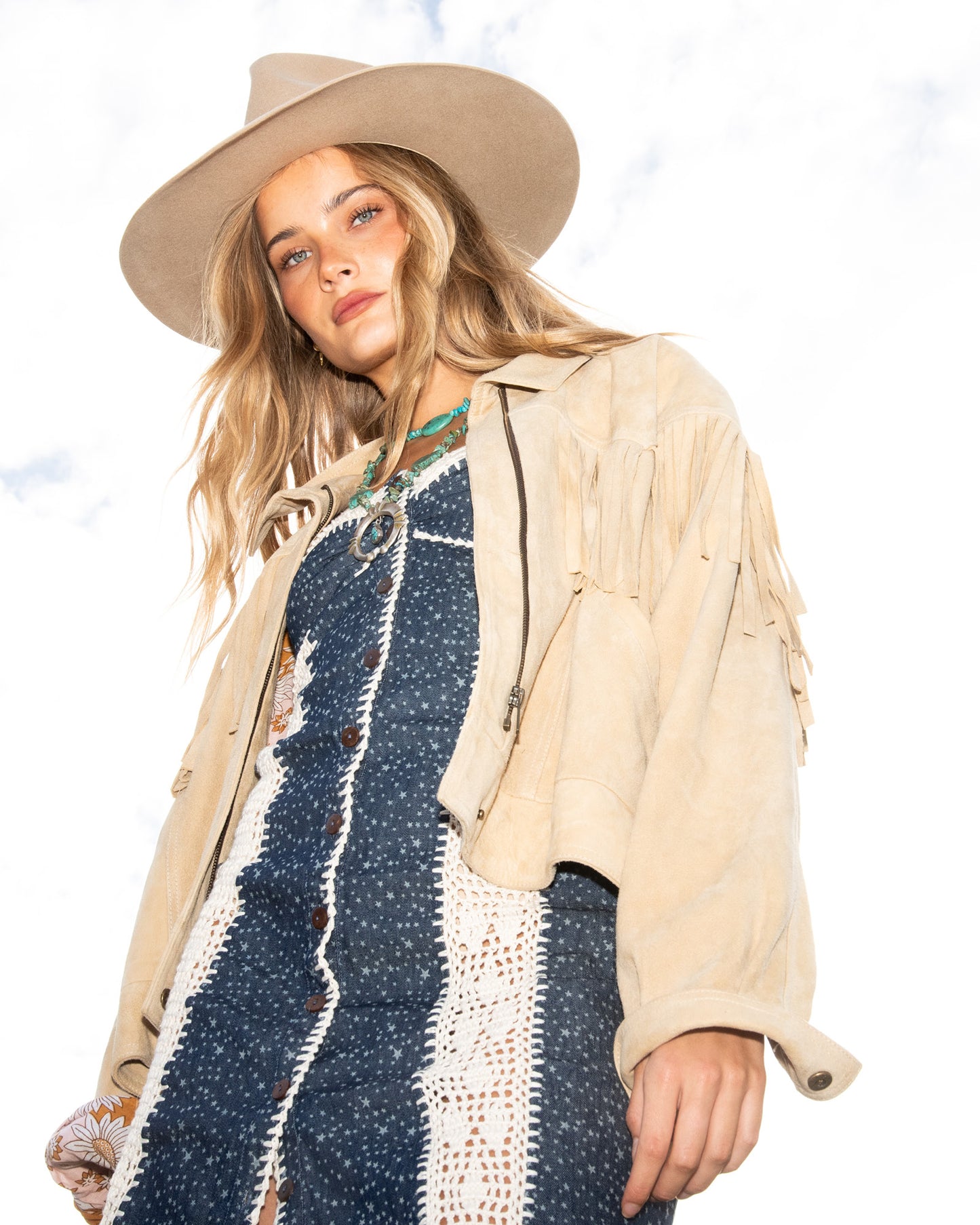 Woman wearing a beige fringe jacket, blue patterned dress, and beige hat against a white background Model wearing The Jose felt hat in natural light

