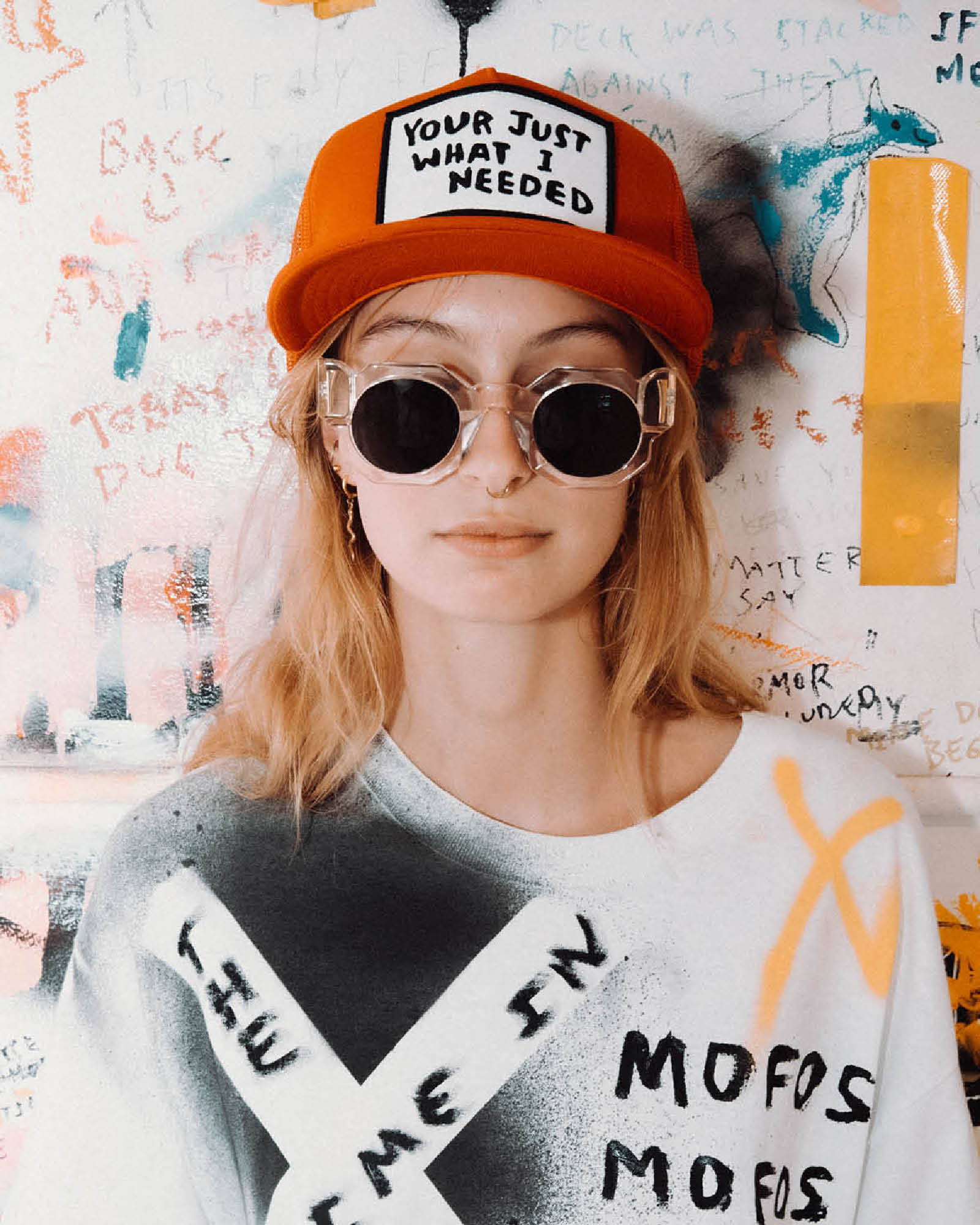 Model wearing red Marty Baptist cap with sunglasses and printed tee in art-covered room.
