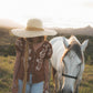 Back view of a child in The Lover Straw Hat walking beside a horse in glowing afternoon light. A poetic image of carefree adventure and natural charm.

