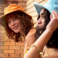 Models wearing FallenBROKENstreet Saturday Bucket Hats in Tangerine and Blue. Cotton chenille wide brim hats with UPF 50+ rating, lightweight and travel-ready. Photographed outdoors, styled casually with bright dresses, showcasing playful unisex summer design.