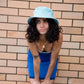 Model wearing Saturday Bucket Hat – Blue by FallenBROKENstreet. Cotton chenille bucket hat with wide brim, cotton inner band, and UPF 50+ sun protection. Styled in a blue slip dress, highlighting casual summer elegance.