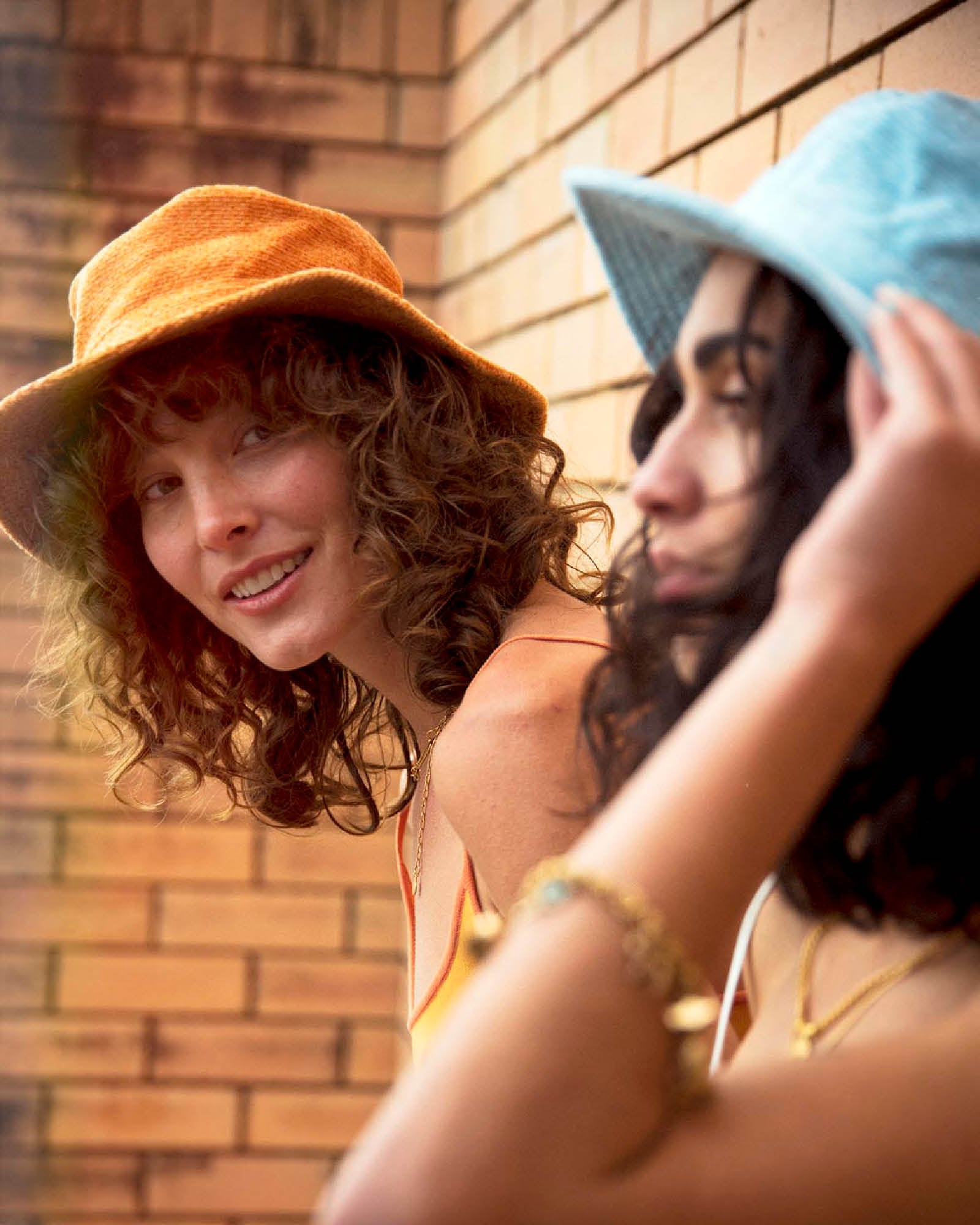 Two models wearing FallenBROKENstreet Saturday Bucket Hats, one in Tangerine and one in Blue. Cotton chenille hats with wide brims, UPF 50+ sun protection, and cotton inner bands. Styled casually outdoors for a playful, unisex summer look perfect for beach days, festivals, and travel.
