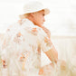 Model wearing the Saturday Bucket Hat – White by FallenBROKENstreet. Cotton chenille wide brim hat with UPF 50+ sun protection, styled with a floral shirt and bright shorts. Photographed outdoors at the beach, highlighting casual unisex summer styling.