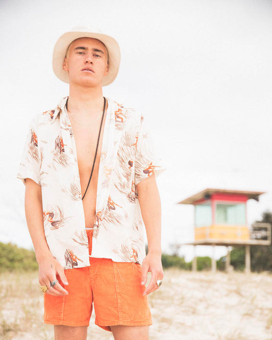 Full-body portrait of model wearing Saturday Bucket Hat – White by FallenBROKENstreet. Lightweight cotton chenille hat with wide brim and UPF 50+ protection. Styled casually with floral shirt and shorts, photographed by the coast, emphasising its travel and summer-ready versatility.