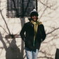 A stylish winter streetwear scene featuring a model wearing The Font Beanie by FallenBROKENstreet — a soft knit beanie with a two-tone pom pom. The model stands casually against a pale wall with winter shadows, dressed in a black coat, yellow scarf, and light denim jeans. This cozy unisex beanie brings playful warmth and urban charm to cold days, ideal for layering with seasonal outerwear.

