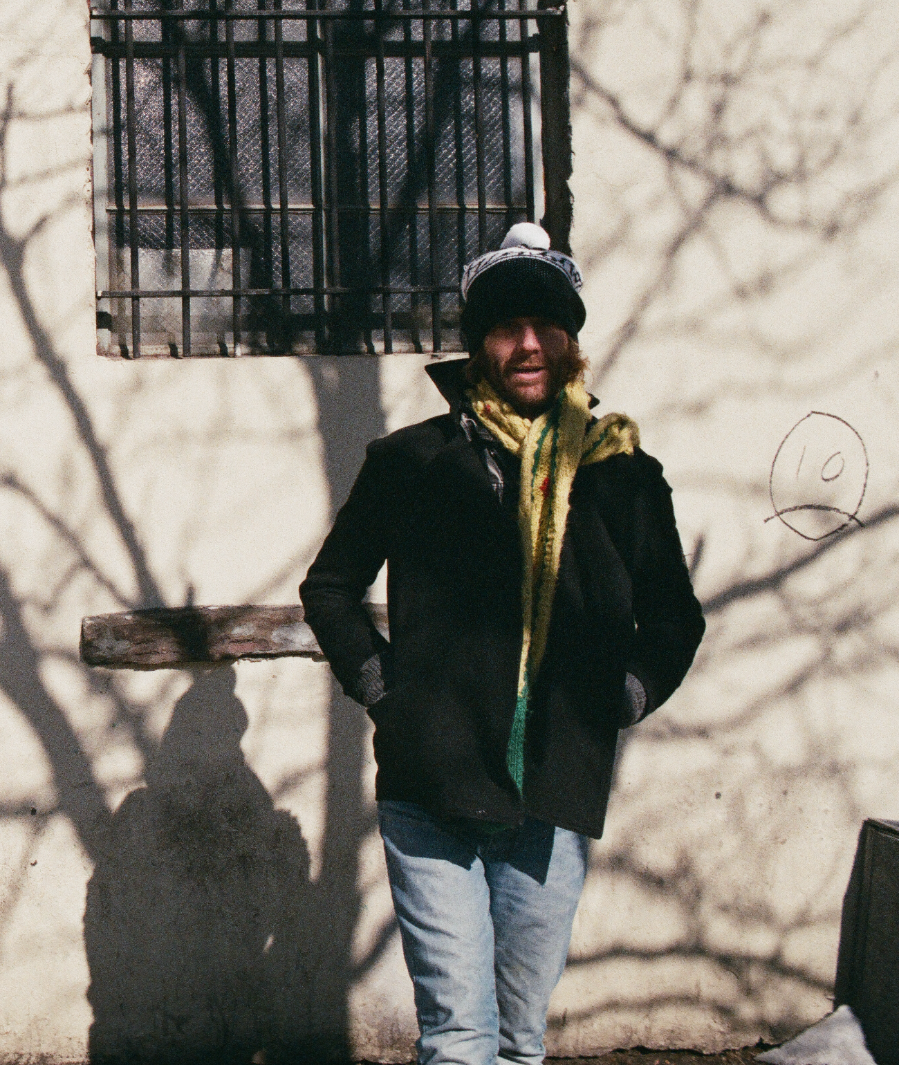 A stylish winter streetwear scene featuring a model wearing The Font Beanie by FallenBROKENstreet — a soft knit beanie with a two-tone pom pom. The model stands casually against a pale wall with winter shadows, dressed in a black coat, yellow scarf, and light denim jeans. This cozy unisex beanie brings playful warmth and urban charm to cold days, ideal for layering with seasonal outerwear.

