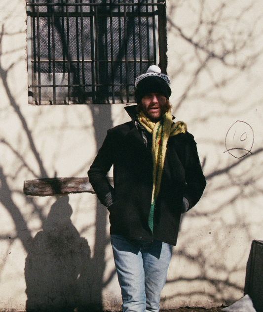 A stylish winter streetwear scene featuring a model wearing The Font Beanie by FallenBROKENstreet — a soft knit beanie with a two-tone pom pom. The model stands casually against a pale wall with winter shadows, dressed in a black coat, yellow scarf, and light denim jeans. This cozy unisex beanie brings playful warmth and urban charm to cold days, ideal for layering with seasonal outerwear.


