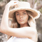 A closer shot of the model adjusting The Seeker Straw Hat, focusing on the frayed edge and the raw organic texture of the raffia straw.