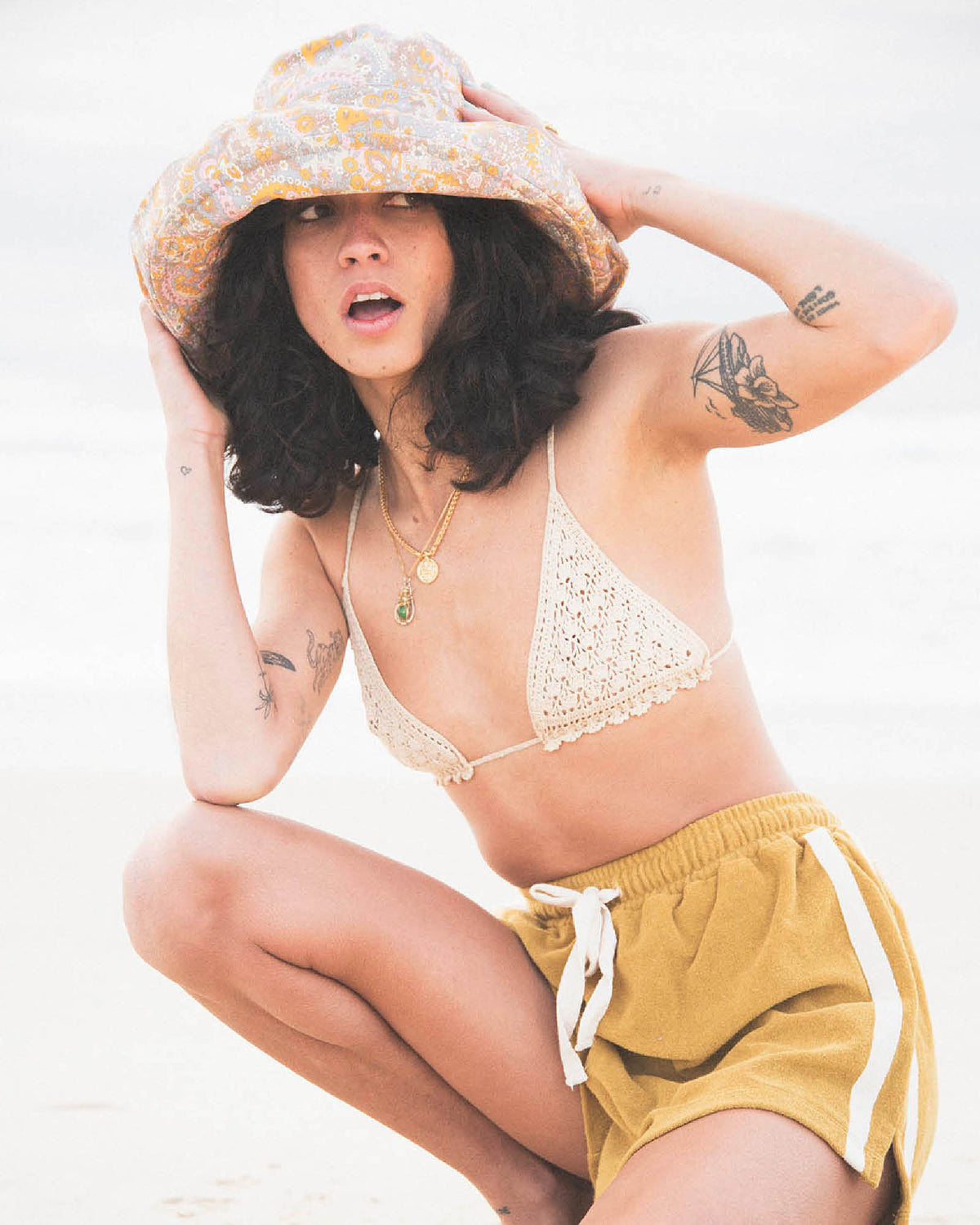 Model wearing the Sunday Bucket Hat – Paisley by FallenBROKENstreet. Lightweight cotton canvas bucket hat with wide brim and all-over paisley print. Styled with a crochet bikini top and shorts at the beach, highlighting its unisex, travel-ready design made for sun protection and coastal adventures.