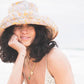Lifestyle photo of the Sunday Bucket Hat – Paisley by FallenBROKENstreet. Lightweight cotton canvas bucket hat with paisley print and wide brim for sun protection. Styled casually with swimwear, photographed at the beach, capturing its playful, summer-ready look and effortless comfort.