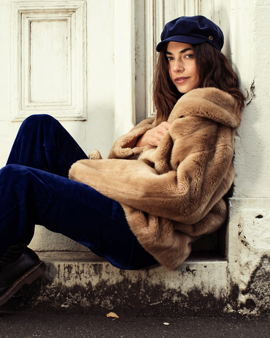 Woman wearing a fur coat and navy outfit sitting against a wall.
