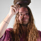 Man with long hair wearing a brown cap against a plain background