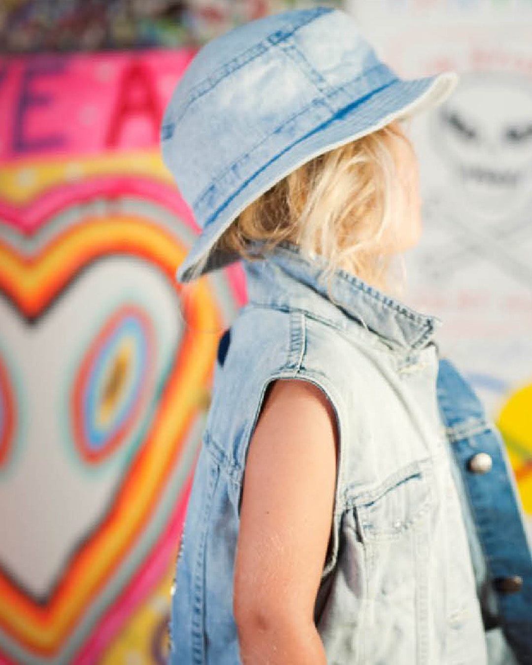Child wearing a denim hat and vest in front of a colorful abstract background
