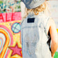 Person wearing a denim vest with rhinestone details and a cap, standing in front of a colorful graffiti wall.