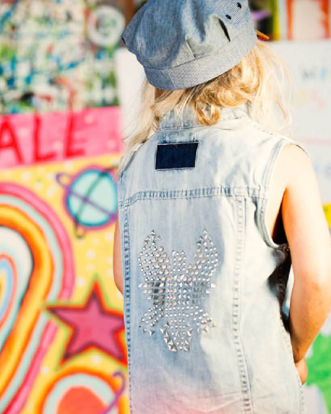 Person wearing a denim vest with rhinestone details and a cap, standing in front of a colorful graffiti wall.