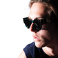 Male model wearing THE COSMOS – Galaxy/Black sunglasses, slightly turned with visible cheekbone and eye contact with camera. Black lenses and cosmic frame styling shown clearly.

