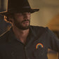 Man wearing a dark hat and shirt with a rainbow patch, standing against a blurred background.