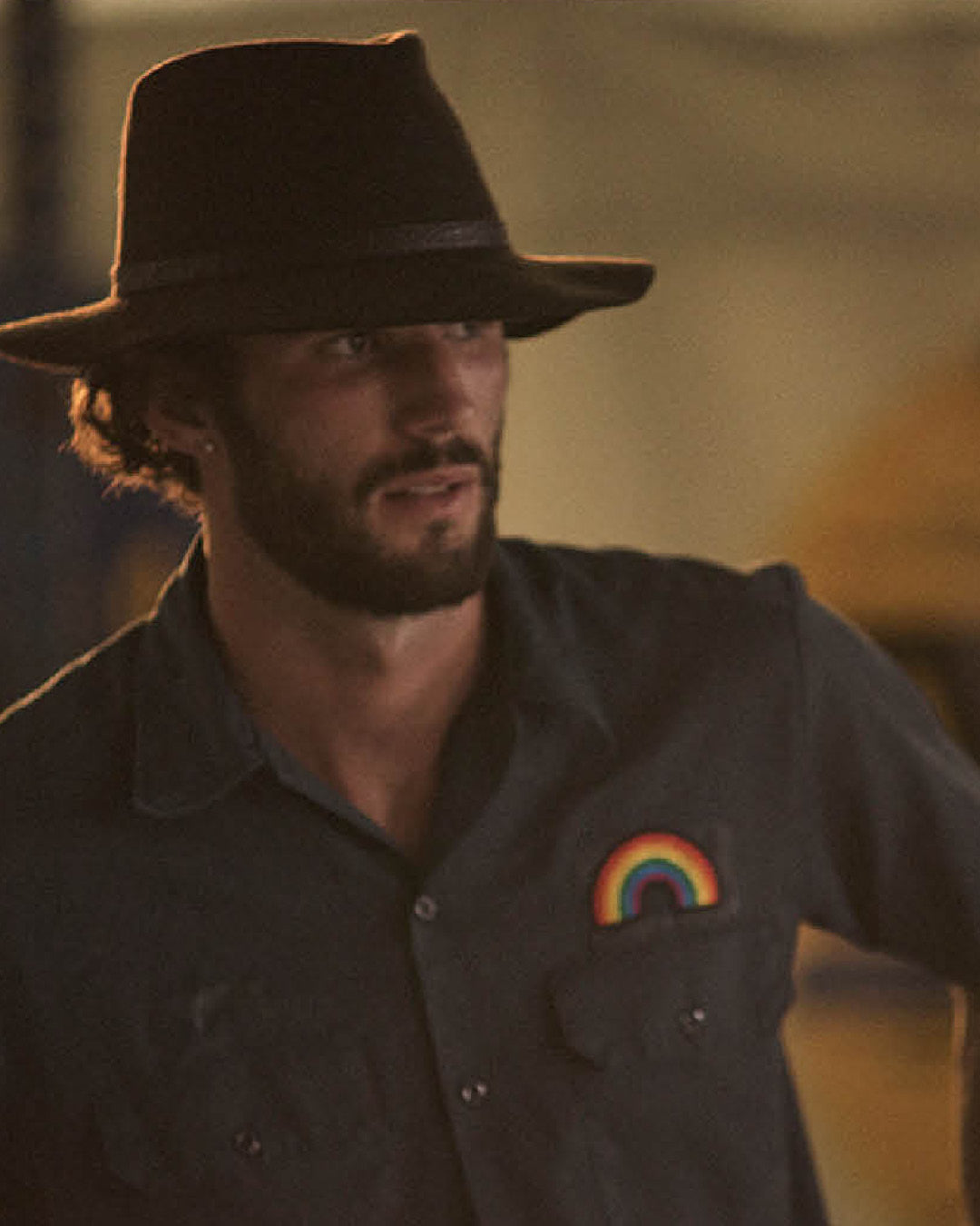 Man wearing a dark hat and shirt with a rainbow patch, standing against a blurred background.